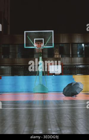 Campo da basket abbandonato di notte con ombrello solitario sul pavimento bagnato, atmosfera urbana tranquilla Foto Stock