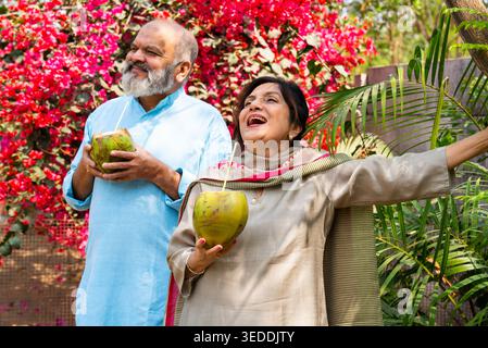 Coppia senior indiana che ha acqua fresca di cocco insieme nella natura seduto nel giardino di casa, bevendo con paglia di verde nariyal e godendo della pace Foto Stock