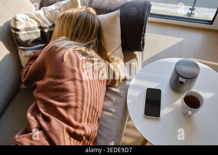 Donna sdraiata a faccia in giù su un divano mentre riposa vicino a un tavolino con una teiera, uno smartphone e una tazza di tè in una stanza soleggiata Foto Stock