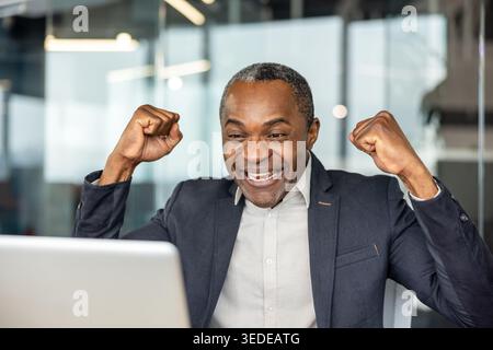 Uomo d'affari maturo che prova gioia e vittoria mentre lavora sul suo portatile in un ufficio moderno, celebrando un successo con pugni alzati e un'espressione allegra Foto Stock
