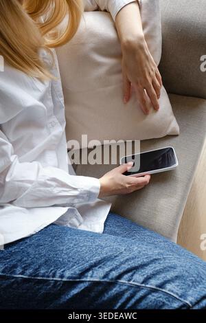 Donna con camicia bianca e jeans seduti su un divano beige con un cuscino e in mano uno smartphone Foto Stock