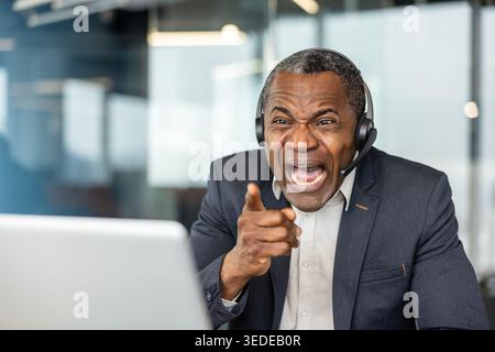 Un uomo frustrato che indossa un auricolare urlando e puntando il dito mentre sta facendo una conversazione online stressante o discute durante una riunione virtuale in un ufficio moderno Foto Stock