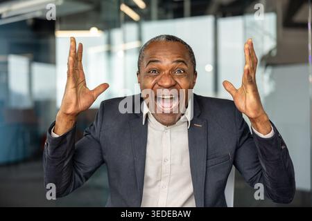 Uomo d'affari maturo che prova gioia e trionfo, festeggia felicemente un risultato significativo o raggiunge un obiettivo aziendale, esprimendo al contempo forti emozioni positive in un ambiente di ufficio Foto Stock