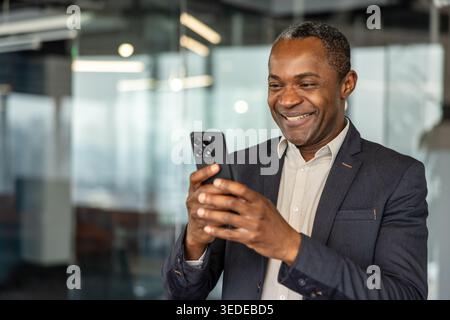 Uomo professionale maturo e felice sorridente mentre tiene in mano uno smartphone, guarda lo schermo e goditi la comunicazione mobile in un ambiente d'ufficio moderno Foto Stock