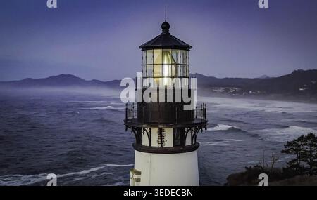 Vista aerea del faro di Yaquina Head con la sua lanterna che si illumina al crepuscolo e si affaccia sulle onde che si infrangono nell'Oceano Pacifico e sulle nebbiose colline costiere ne Foto Stock