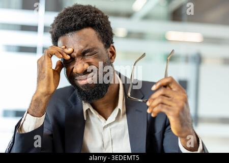 Giovane imprenditore stressato che chiude gli occhi, tenendo la fronte con una mano e gli occhiali con l'altra, soffre di un forte mal di testa e di un eccessivo lavoro in un ambiente di ufficio moderno Foto Stock
