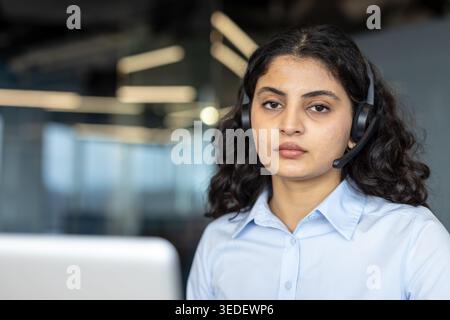 Giovane donna che guarda direttamente la fotocamera, lavora in un ufficio moderno, indossa una cuffia professionale, pronta a fornire assistenza clienti e assistenza tecnica Foto Stock