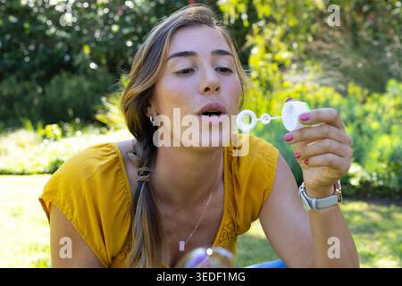 Donna inclinata in avanti in giardino con indosso una camicetta gialla, che soffia la bolla con la bacchetta, bottiglia bianca Foto Stock
