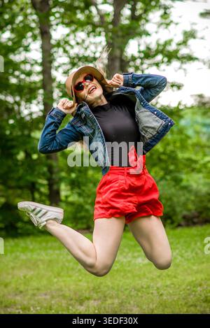 Ragazza con cappello di paglia, giacca in denim, pantaloncini rossi e sneaker bianche che indossano occhiali da sole a forma di cuore rosso e rossetto rosso brillante che salta. Foto Stock
