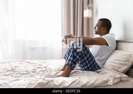 Uomo nero seduto sul letto che guarda la finestra con cura in una camera da letto luminosa Foto Stock