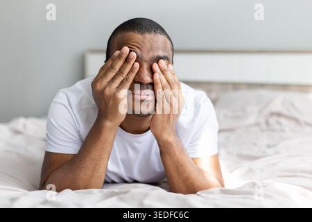 Uomo afroamericano disteso sul letto che copre il viso con le mani, esprimendo stress Foto Stock