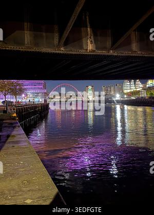 Vista notturna del Millennium Bridge che attraversa il fiume Tyne a Newcastle upon Tyne con vivaci luci viola della città riflesse nell'acqua Foto Stock