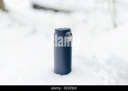 Matraccio thermos blu scuro in posizione verticale sulla neve fresca in una fredda giornata invernale all'aperto. La bottiglia isolata si alza sopra il terreno ghiacciato e pronta a servire bevande calde. Attrezzatura essenziale per le escursioni invernali Foto Stock