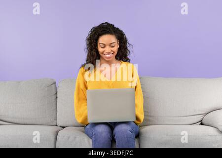 La donna si siede su un divano con un notebook e sorride mentre guarda lo schermo Foto Stock