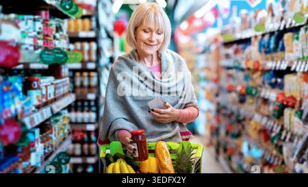 Donna anziana che fa acquisti di generi alimentari nella corsia del supermercato con prodotti colorati intorno a lei Foto Stock
