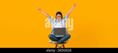 Una giovane donna si siede su uno sgabello con un laptop sul giro e solleva le braccia con entusiasmo Foto Stock