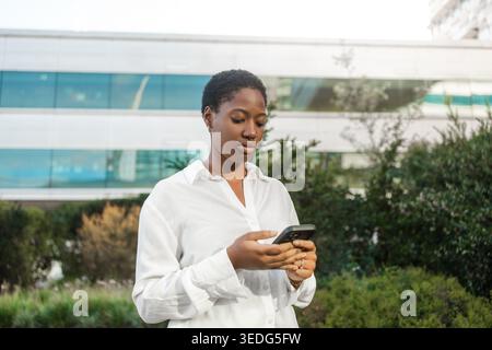Giovane donna d'affari in camicia bianca che usa lo smartphone mentre si trova vicino a un moderno edificio per uffici durante il giorno Foto Stock