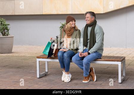 Coppia senior seduta sulla panchina in piazza lastricata con borse e borsetta, con sciarpe Foto Stock