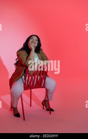 Donna di mezza età seduta su una sedia a doghe in metallo rosso in studio, appoggiata, sorridente, con anello Foto Stock