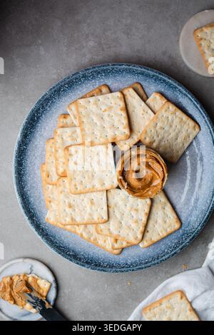 Vista dall'alto del burro di arachidi e dei cracker su un piatto blu, vista dall'alto di un vaso di burro di arachidi con cracker croccanti su un piano di lavoro grigio Foto Stock