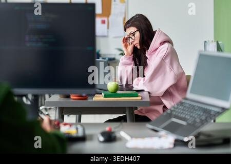 Giovane donna caucasica adulta che lavora su un notebook in ufficio, tocca gli occhiali e si concentra sull'attività di codifica, rappresentando un flusso di lavoro tecnologico mirato. Per lo sviluppo di software o il marketing di carriera IT Foto Stock