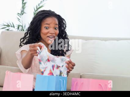 Donna afroamericana seduta su un divano beige con piano floreale e borse per lo shopping blu e rosa nelle vicinanze. Indumenti, moda, pastello, pianta, casa, accogliente, ricci Foto Stock