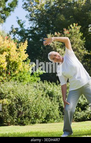 Donna anziana che pratica stretch laterale su un prato erboso con arbusti, T-shirt bianca grigia, spazio copia. Anziani, esterni, fitness, benessere, natura, garde Foto Stock