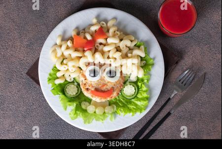 Cibo divertente per bambini - ragazza con viso sorridente fatta di cutlet di tacchino, pasta cavatappi, olive, pepe, uovo e cetriolo su piatto bianco Foto Stock
