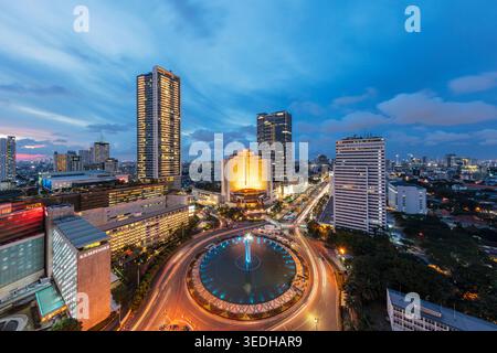 Vista ultra-ampia dell'ora blu di Bundaran HI e Selamat Datang Monument con percorsi dinamici a semaforo da un tetto a Giacarta, Indonesia Foto Stock