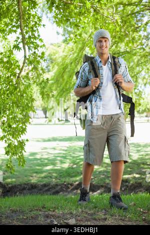 Uomo che indossa berretto in maglia e pantaloncini cargo, in piedi in un parco illuminato dal sole e porta con sé uno zaino con tappetino arrotolato. Escursioni, zaino, Sleepingpad, baldacchino, spazio verde, Foto Stock