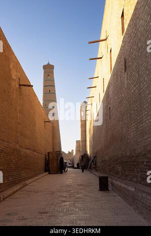 Vicolo stretto di Ichan Kala che conduce al minareto Islam Khodja a Khiva, Uzbekistan. Una calda luce dorata illumina le antiche pareti di mattoni al tramonto. Ideale per pubblicazioni di viaggio e per il patrimonio culturale... Foto Stock