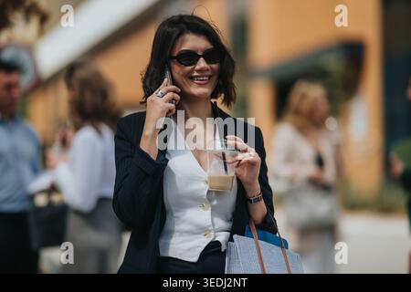 Donna d'affari sorridente al telefono con caffè ghiacciato all'aperto tra colleghi in una riunione urbana informale Foto Stock