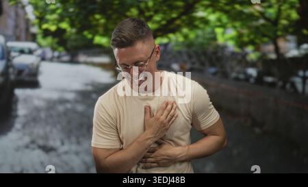Uomo che ride con la mano sul petto e tiene lo stomaco, sorride per strada mentre indossa occhiali e camicia beige; sollievo gratitudine. Foto Stock