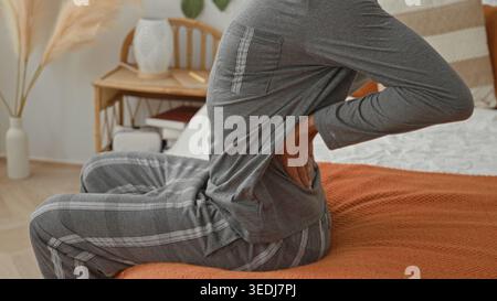 Uomo seduto sul letto che tiene la parte inferiore della schiena con la mano, indossa pigiami grigi su una coperta a maglia arancione in una camera da letto dell'edificio con comodino e lampada visibili; p Foto Stock