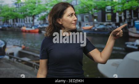 Donna ispanica di mezza età punta il dito verso destra, occhi fissi, in piedi vicino a barche ormeggiate e edifici in mattoni su una strada con maniche corte navy Foto Stock