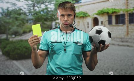 L'arbitro uomo tiene la carta gialla nella mano sinistra e la palla da calcio a destra, indossando fischio e cuffie su strade acciottolate dopo edifici di pietra mentre cammina Foto Stock