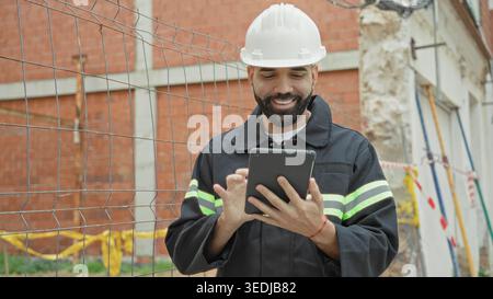 Man Architect in elmetto e giubbotto riflettente punta le dita sul tablet e tocca lo schermo della recinzione del cantiere, pianificando la sicurezza. Foto Stock