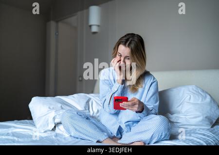 Mattina online con il telefono. Una donna contenta usa lo smartphone seduto a letto scorrendo le ragnatele dopo il risveglio Foto Stock