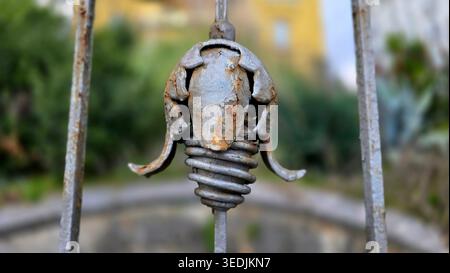 Una foto dettagliata di un ornamento metallico grigio con forme a spirale e floreali su un vecchio cancello. Lo sfondo presenta una morbida vegetazione sfocata Foto Stock