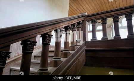 Una vista ad angolo basso di una grande e polverosa scalinata in legno in un edificio storico abbandonato. Il banister presenta colonne decorative intagliate Foto Stock