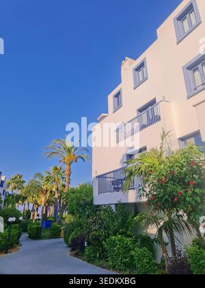 Luminoso edificio di appartamenti in stile mediterraneo bianco con balconi, palme tropicali, lussureggiante giardino paesaggistico, passerella e cielo azzurro limpido in una giornata di sole. Foto Stock