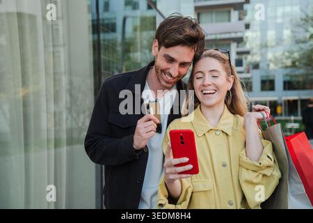 Joyful Couples condivide un'esperienza di shopping piacevole mentre controllano sul proprio smartphone offerte e promozioni interessanti, migliorando il loro retail Foto Stock