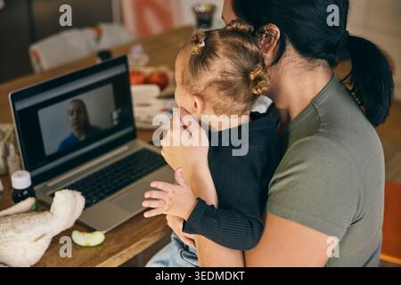 Vista ad angolo alto della madre che spiega la febbre della figlia al medico durante una videochiamata dal computer portatile mentre è seduto a casa Foto Stock