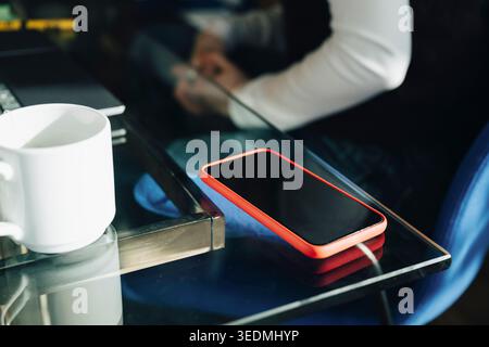 Smartphone con schermo vuoto accanto alla tazza da caffè sulla scrivania in vetro dell'ufficio Foto Stock