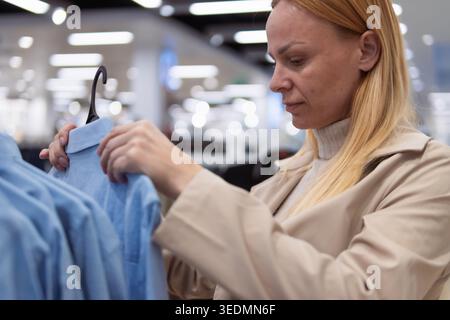 Donna che sfoggia camicie blu su grucce in un moderno negozio di abbigliamento, concentrandosi sulla sua esperienza di acquisto e selezionando nuovi capi di abbigliamento per il suo stile personale Foto Stock