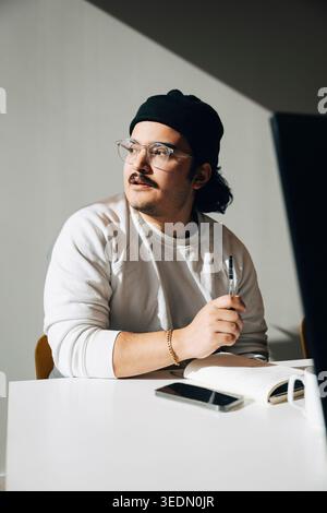 Giovane uomo d'affari attento che guarda lontano mentre prepara appunti in diario in ufficio Foto Stock