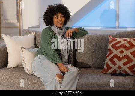 Donna afroamericana seduta su un divano grigio con maglione verde e sciarpa grigia, smartphone nelle vicinanze Foto Stock
