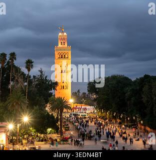 Il minareto della moschea di Koutoubia si illumina al crepuscolo sopra una vivace piazza di Marrakech, con palme, folle, lampioni e nuvole d'atmosfera, trasmettendo alta qualità Foto Stock