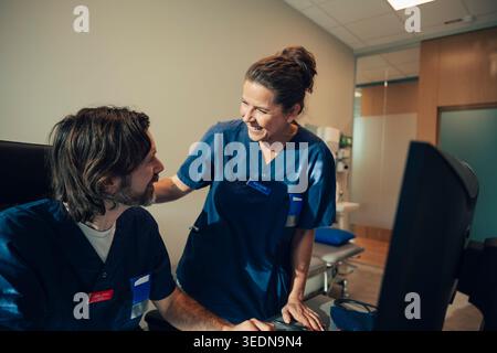 Donna sorridente esperto medico che parla con il collega di sesso maschile seduto alla scrivania in ufficio in ospedale Foto Stock