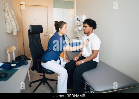 Donna medico professionista che ascolta il battito cardiaco del giovane uomo attraverso lo stetoscopio in sala d'esame all'ospedale Foto Stock